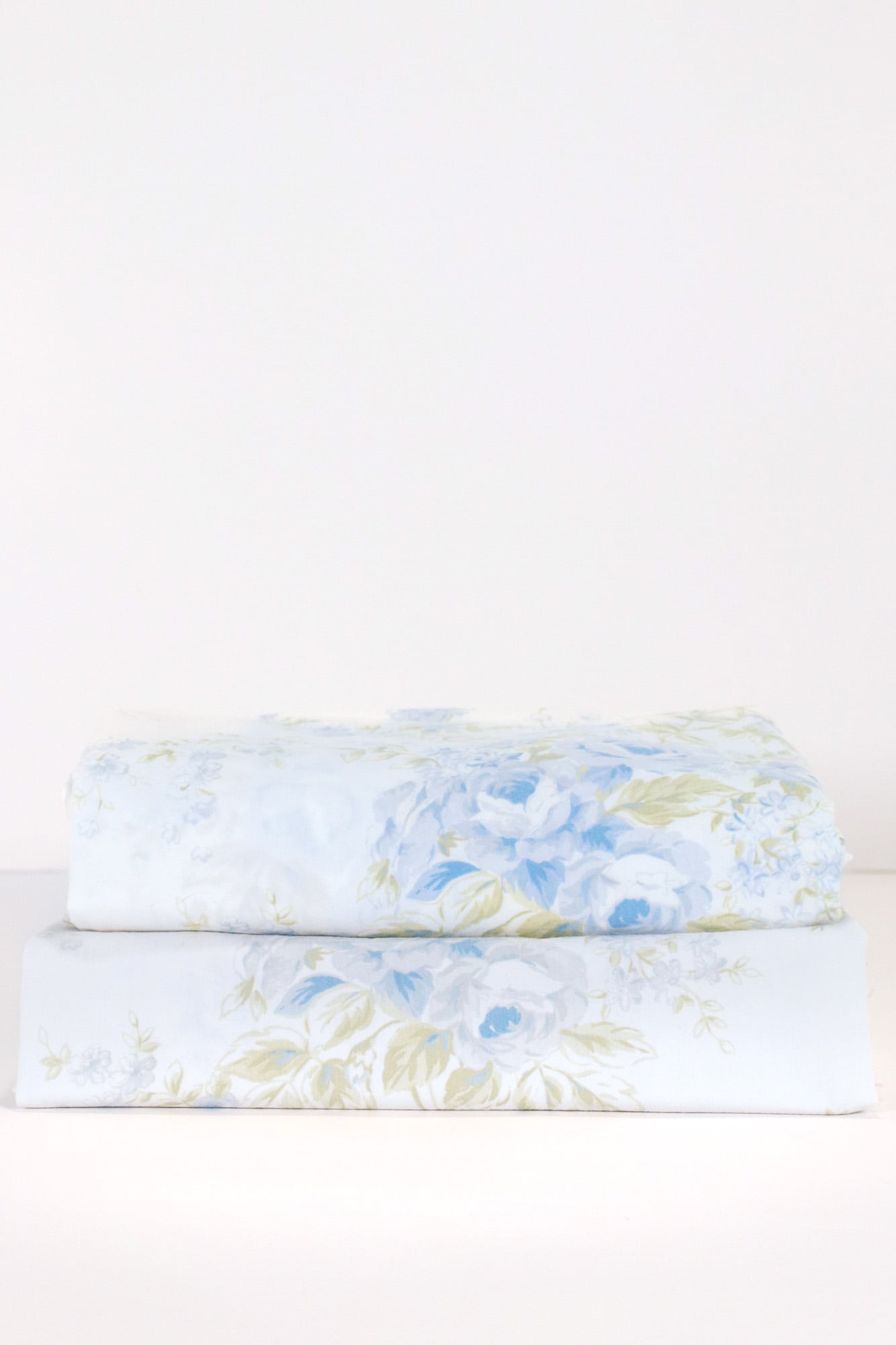 Front view of a neatly folded and stacked pair of Simply Shabby Chic British Rose 36 by 84 inch curtain panels with soft blue floral print on white